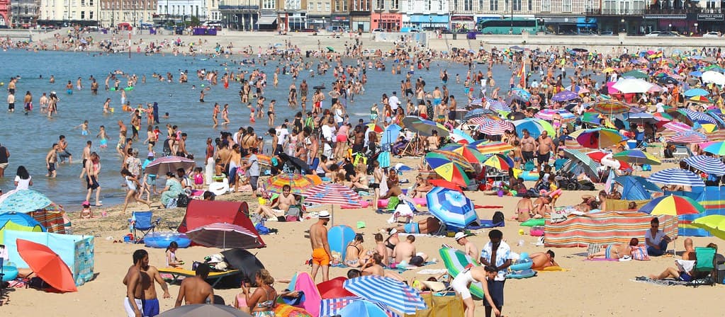 O plajă aglomerată, cu mulți oameni în apă și la soare, sub umbrele și corturi de plajă. (Foto de arhivă: Graham Ó Síodhacháin / BY-SA 2.0, Openverse)