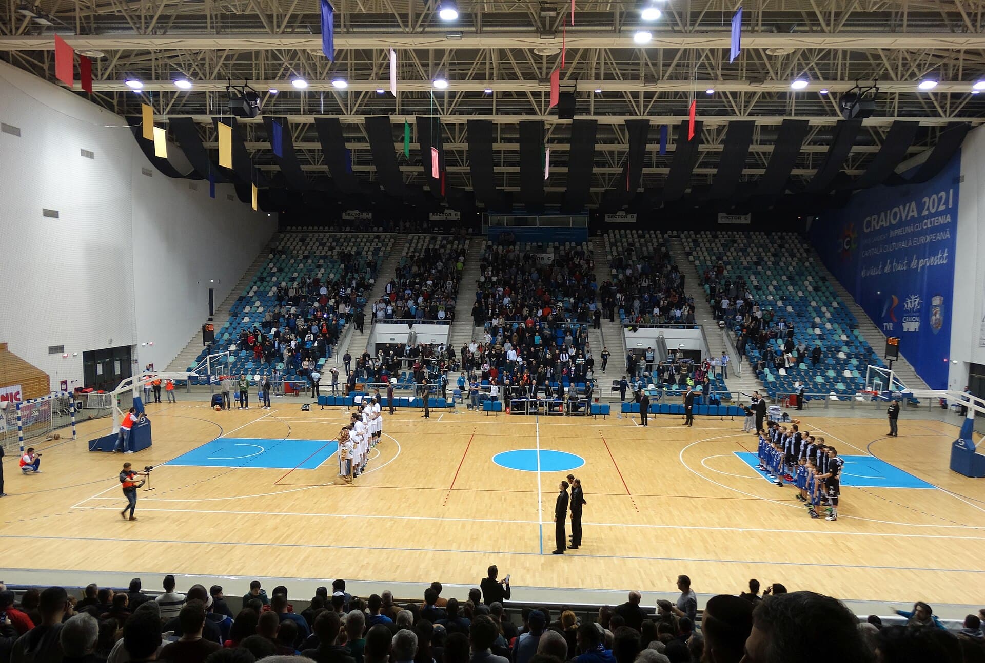 Un meci de baschet juvenil într-o sală, cu două echipe U18 aliniate pe teren și spectatori în tribune. (Foto: Nenea hartia / CC BY-SA 4.0, Wikimedia Commons)