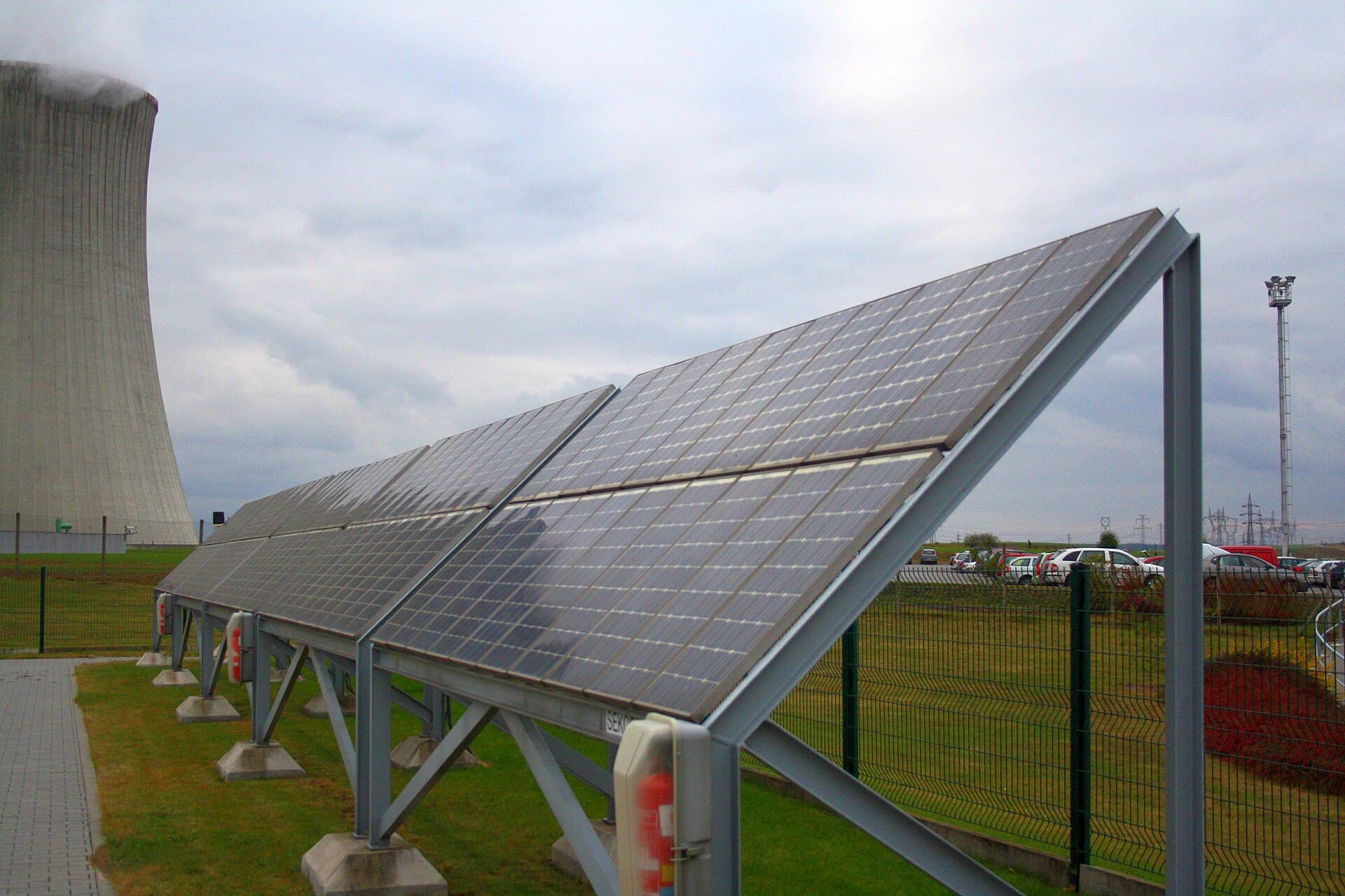 Un șir de panouri solare fotovoltaice montate pe o structură metalică, într-o zonă industrială, lângă un turn de răcire. (Foto de arhivă: Frettie / CC BY 3.0, Wikimedia Commons)
