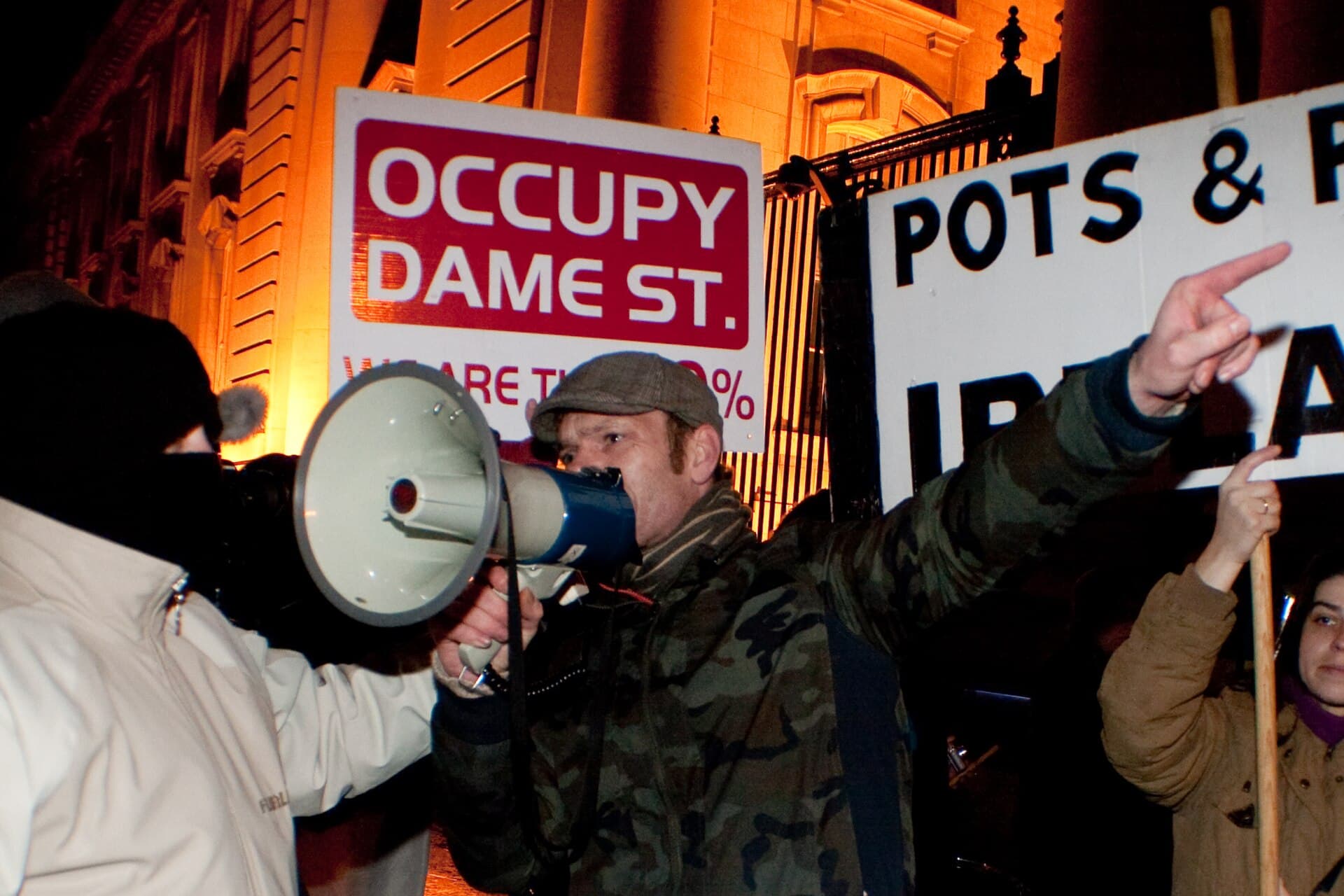 Un protest nocturn, cu manifestanți care țin pancarte și folosesc un megafon în fața unei clădiri. (Foto de arhivă: Paul Reynolds from Dublin, Ireland / CC BY 2.0, Wikimedia Commons)