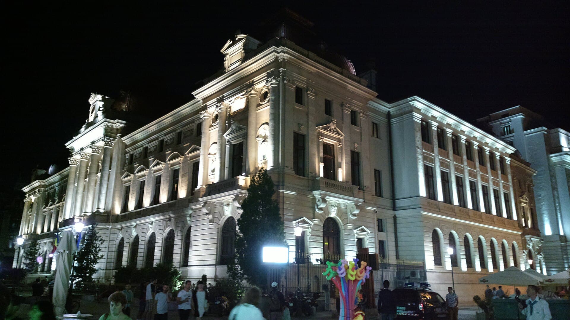 O clădire instituțională impunătoare, iluminată noaptea, într-un spațiu urban din România. (Foto: Korinna / CC BY-SA 3.0 ro, Wikimedia Commons)