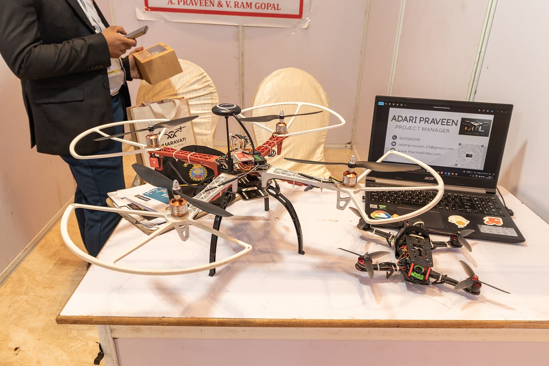 Două drone de tip quadcopter expuse pe o masă, lângă un laptop și un participant parțial vizibil. (Foto de arhivă: iMahesh / CC BY-SA 4.0, Wikimedia Commons)