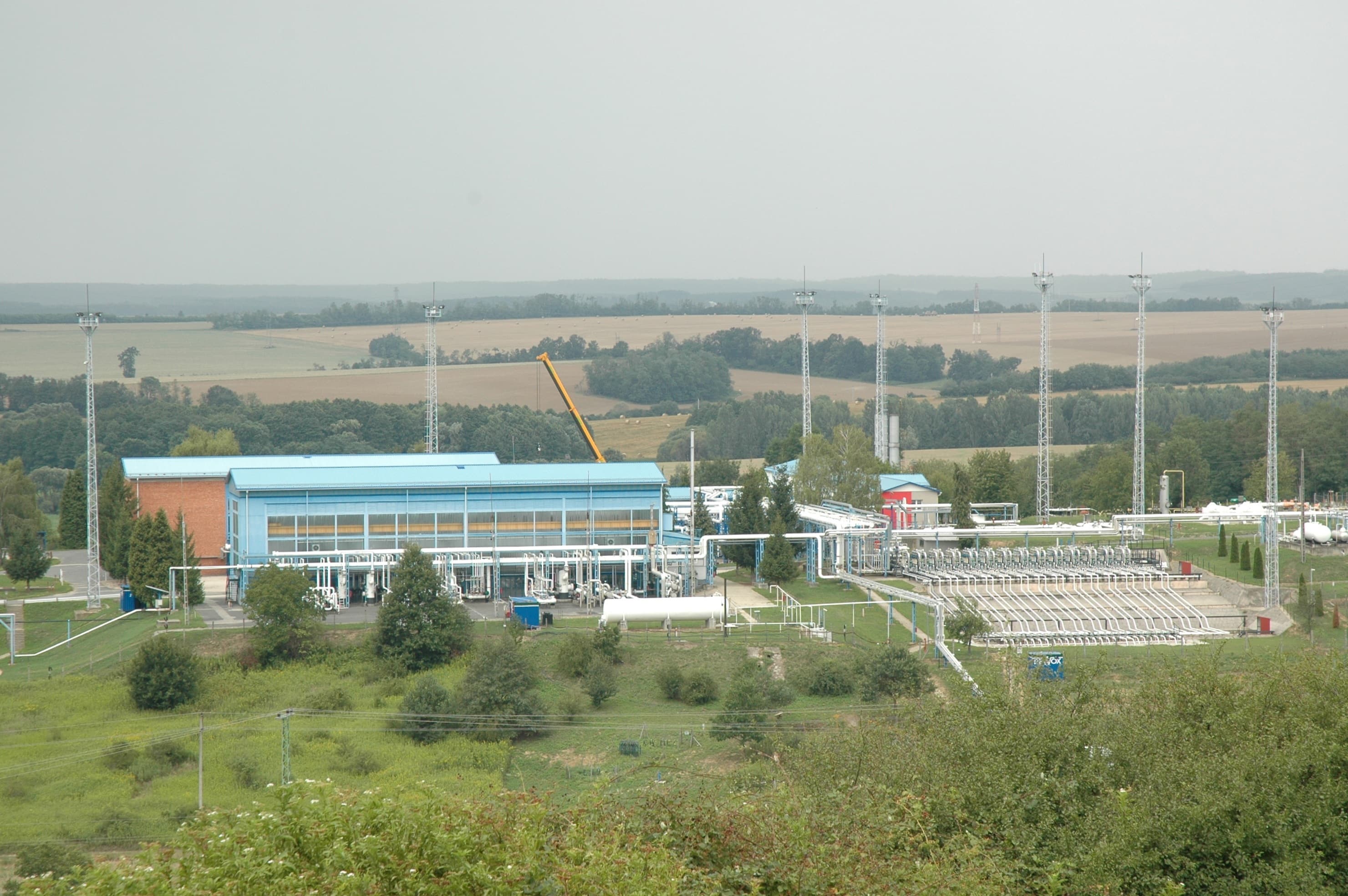 Un complex industrial de infrastructură energetică, cu clădiri, conducte și instalații tehnice, amplasat într-o zonă rurală. (Foto de arhivă: VargaA / BY-SA 3.0, Openverse)