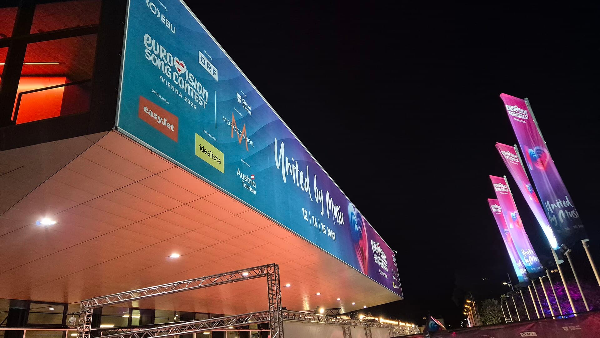 Panouri și bannere oficiale Eurovision Song Contest amplasate pe fațada unei clădiri și de-a lungul unui spațiu exterior, fotografiate noaptea. (Foto: Eurovince / CC BY 4.0, Wikimedia Commons)