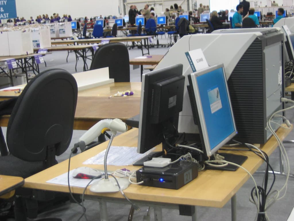 O sală mare cu mese, calculatoare și echipamente de vot, unde mai multe persoane participă la o procedură electorală. (Foto de arhivă: secretlondon123 / BY-SA 2.0, Openverse)