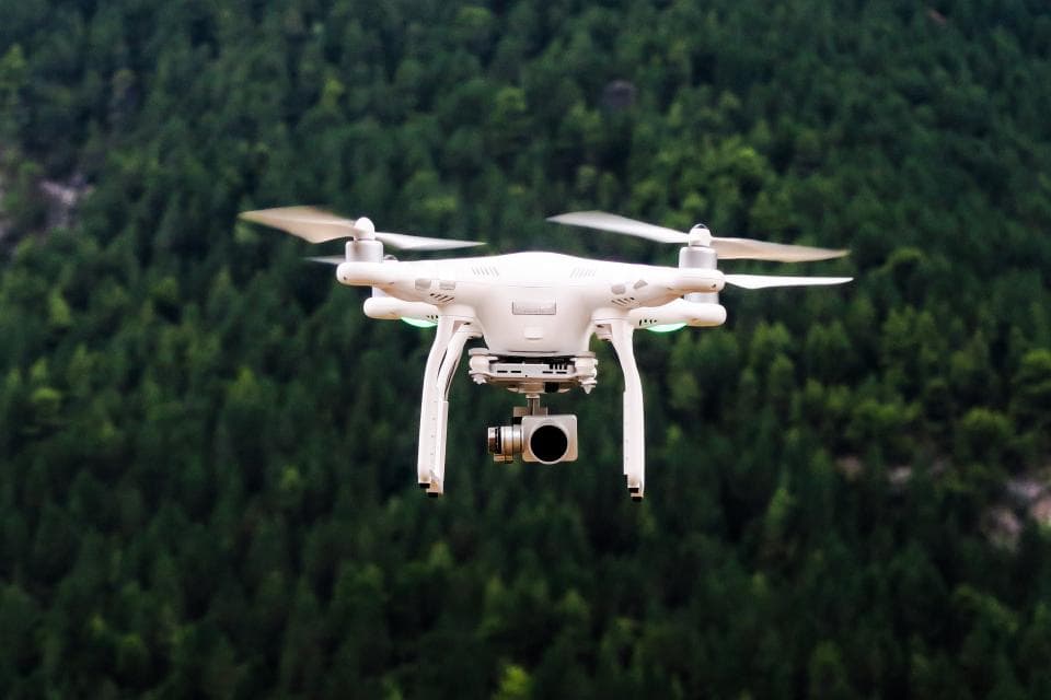 O dronă albă, cu cameră, surprinsă în zbor deasupra unei păduri. (Foto ilustrativă: Jason Blackeye / CC0 1.0, Openverse)