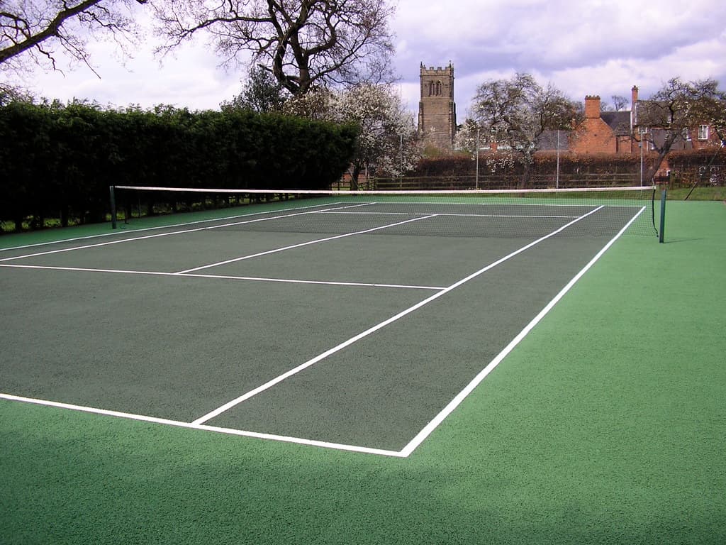 Un teren de tenis în aer liber, fără jucători, într-un parc, cu clădiri și copaci în fundal. (Foto de arhivă: Soft Surfaces Ltd / BY 2.0, Openverse)
