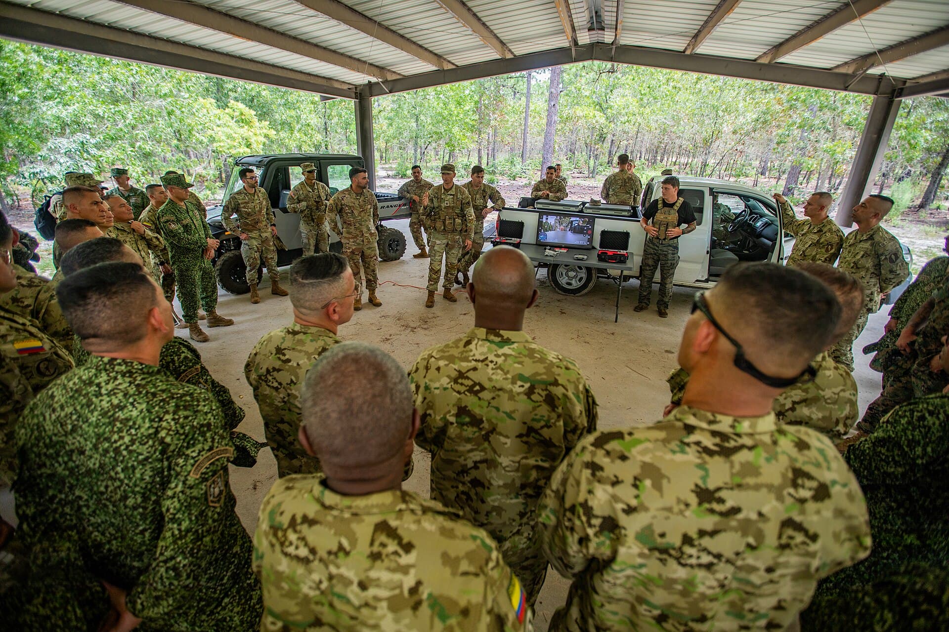 Militari în uniforme de camuflaj reuniți în jurul unui vehicul militar, într-un adăpost acoperit, în timpul unei activități de instruire sau informare. (Foto de arhivă: U.S. Army photo by Sgt. Joshua Taeckens / Public domain, Wikimedia Commons)