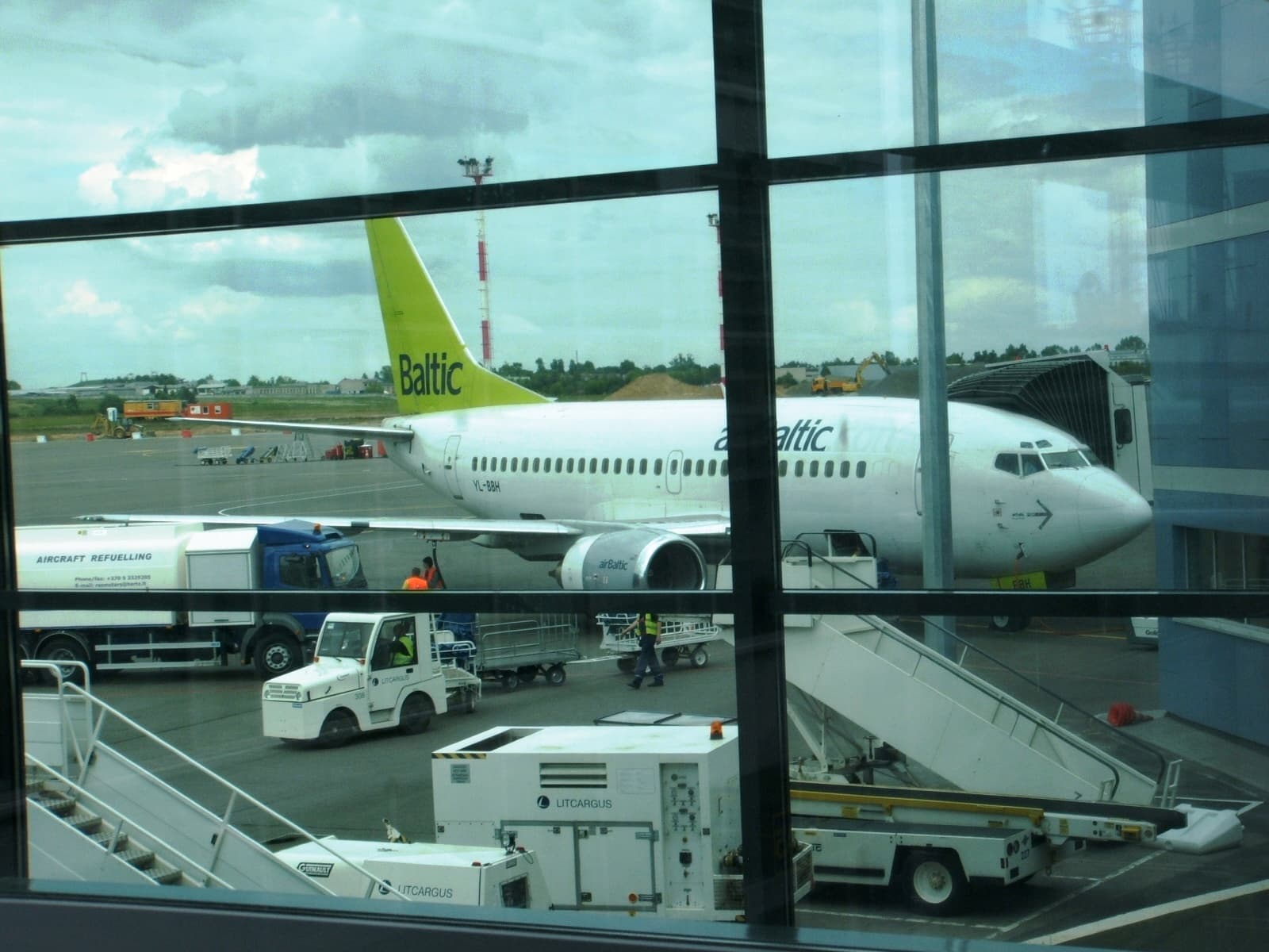 Un avion al companiei airBaltic pe platforma unui aeroport, văzut prin ferestrele terminalului. (Foto: Grobuonis / Public domain, Wikimedia Commons)
